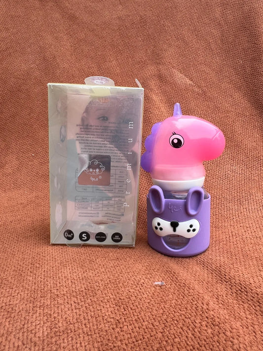 A pink and purple unicorn-shaped baby bottle with a matching purple bunny-shaped holder, shown next to its clear plastic packaging box on a textured brown fabric background.