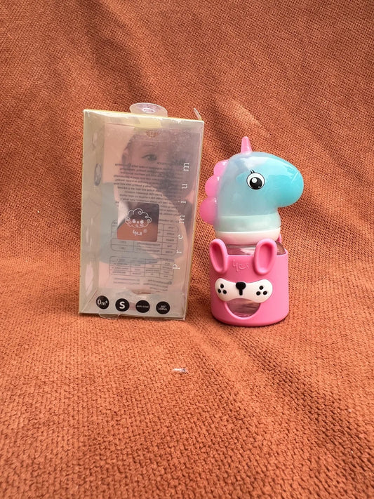 A small pink and light blue unicorn-shaped bottle or container with a cartoon face, shown next to its translucent retail box on a textured orange-brown fabric background.